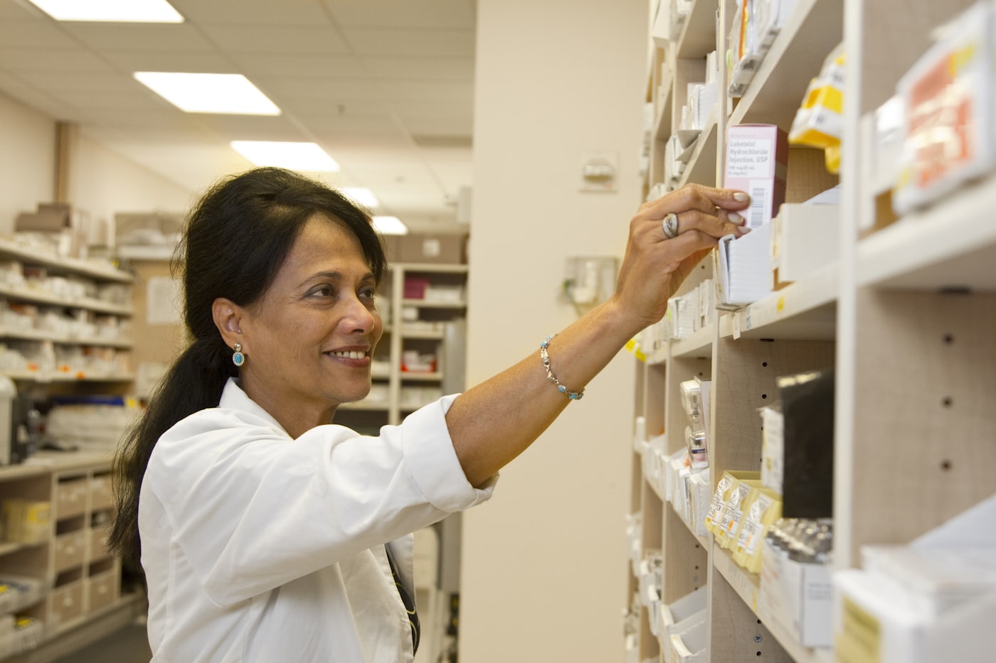 A pharmacist in a white coat reaching for a medicine box on a shelf